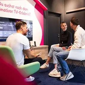 Drei Personen im Büro diskutieren TV-Streaming vor großem Bildschirm.